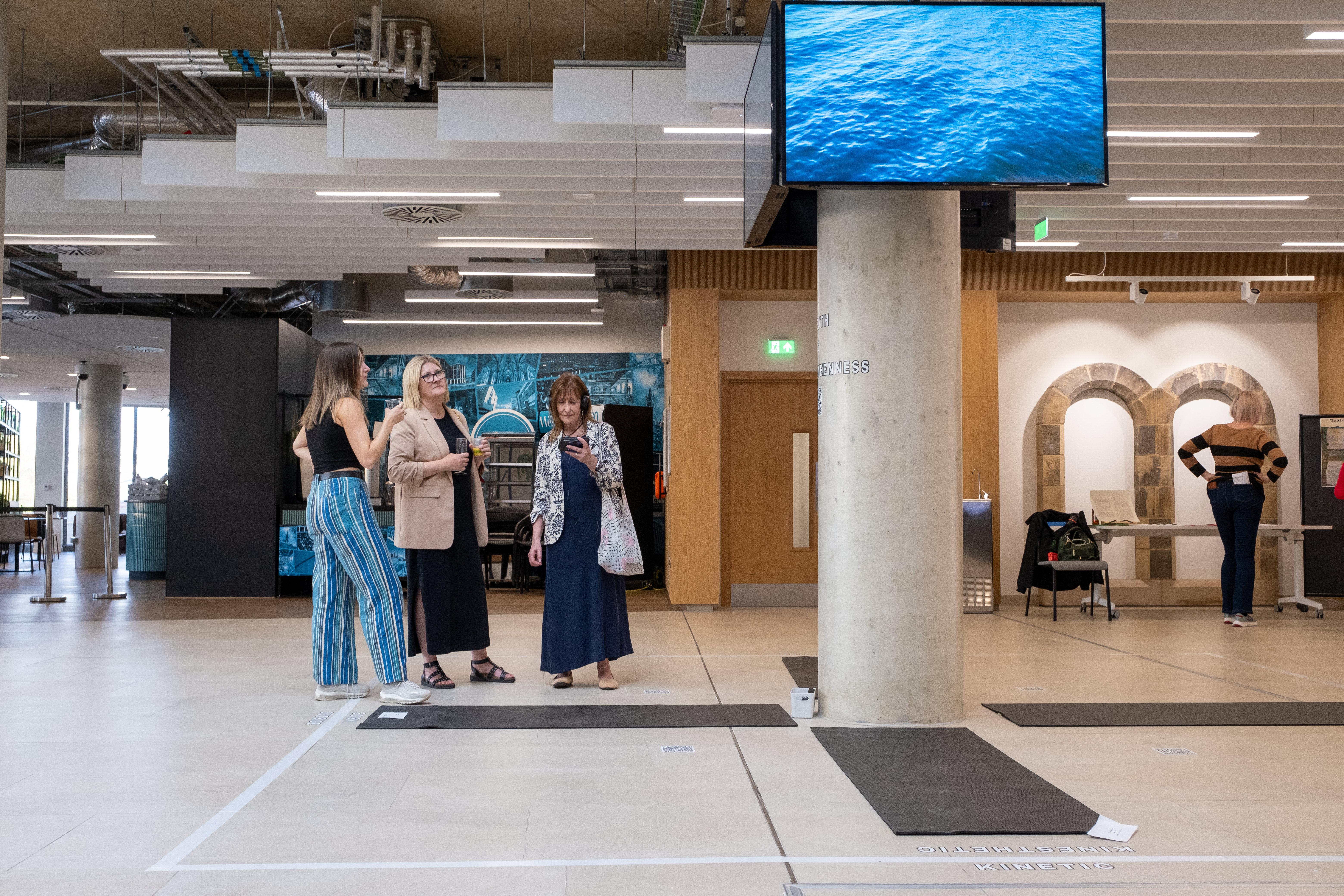 three women look up at screens featuring blue waves on a pillar, four yoga mats are laid out on the floor surrounding the pillar