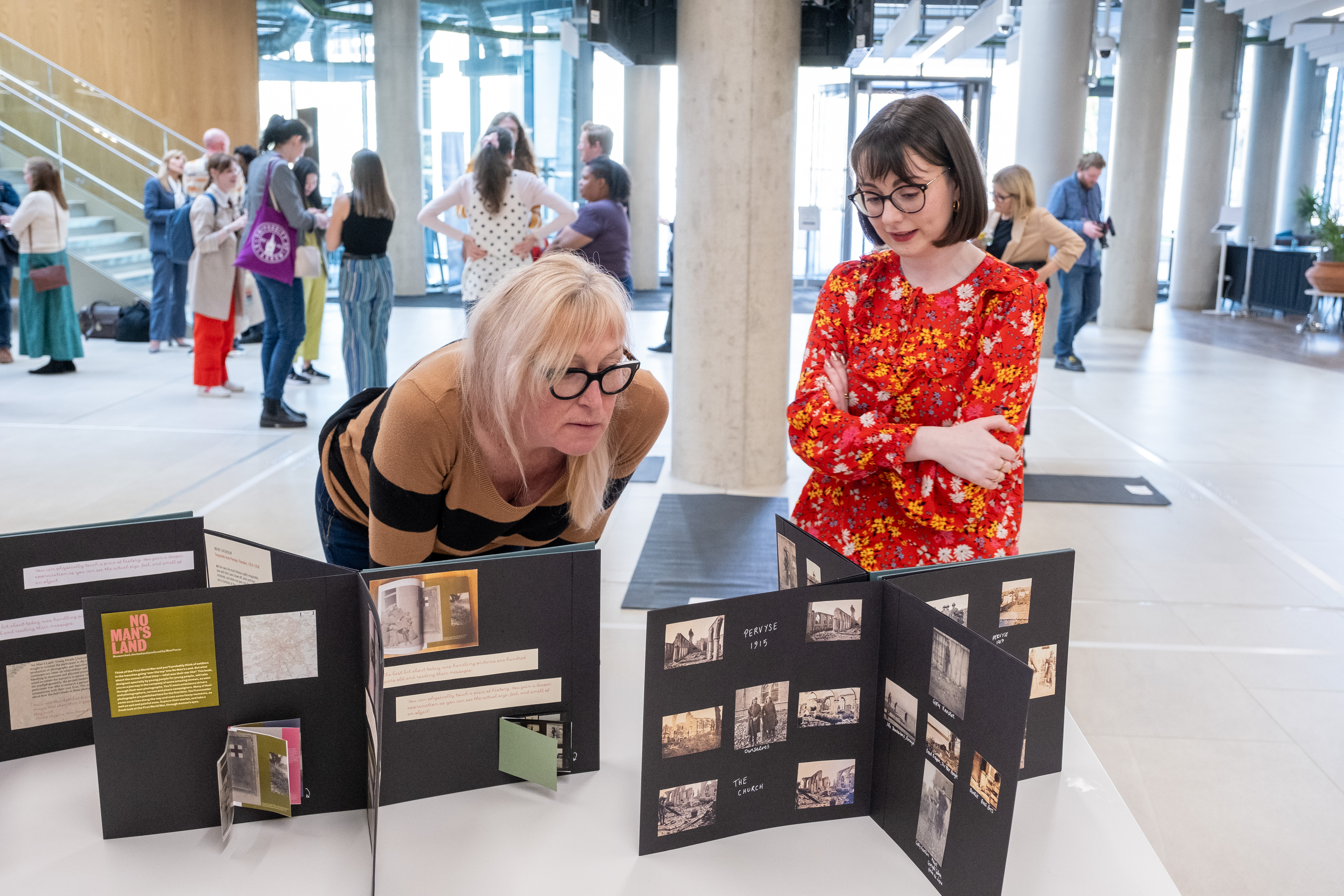 3 book displays sit on a table, a woman crouches down to read them whilst another woman speaks to her