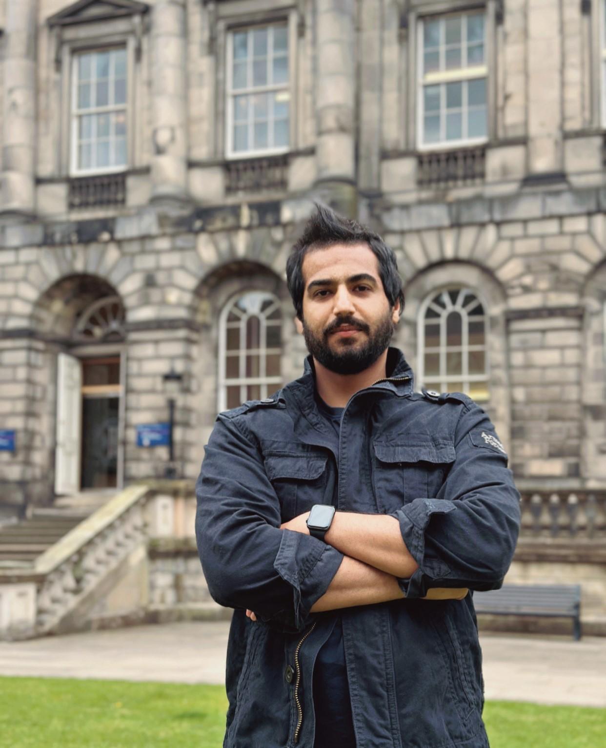 Ahmed H. Alhuwayshil​ Profile Photo - man standing with arms folded outside University building